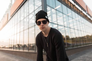 Şık örme siyah şapkalı Amerikalı genç yakışıklı hipster adam şehirde modern bir cam binanın arka planda bir t-shirt moda güneş gözlüğü. Çekici adam model. Erkek giyim.