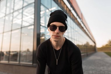 Koyu güneş gözlüğü örme şapka şık siyah gömlek Yakışıklı kentsel genç hipster adam sokakta modern binanın yakınında ayakta poz. İyi adam bir yaz gününde şehirde yürüyor.