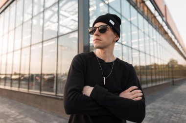 Şehirde modern bir cam binanın arka planda poz bir t-shirt modatrendy güneş gözlüğü şık örme siyah şapka güzel genç yakışıklı hipster adam. Amerikalı çekici adam. Stil.