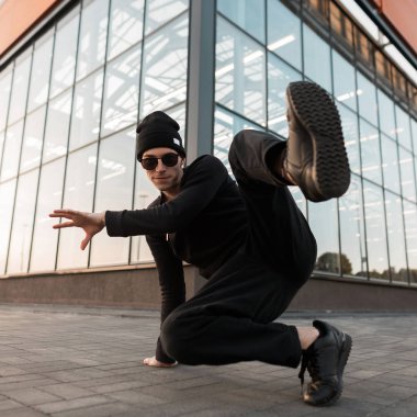 Bir cam binanın yakınında şehirde şık bir gömlek dans break dans güneş gözlüğü spor ayakkabı lı siyah moda örme şapka profesyonel genç adam dansçı. Güzel hipster adam açık havada.