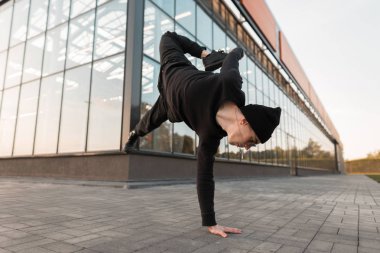 Siyah örgü şapkalı, şık gömlekli, kot pantolonlu genç, yakışıklı hippi şehirde cam bir binanın yanında dans ediyor. Şehir çocuğu break dansçı dışarıda amuda kalkıyor..