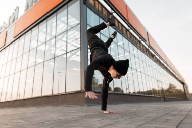 Bir cam binanın yakınındaki sokakta bir örgü şapka dans break dans şık siyah giysiler içinde Modern genç adam dansçı. Şehirde el standı yapan çekici bir adam. Sokak stili. İyi günler.