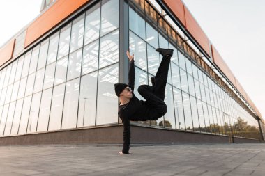 Şık güneş gözlüğü bir örgü şapka siyah şık giysiler modern genç profesyonel erkek dansçı bir cam binanın yakınında bir handstand yapıyor. Yakışıklı hipster adam açık havada dans ediyor. Sokak stili.
