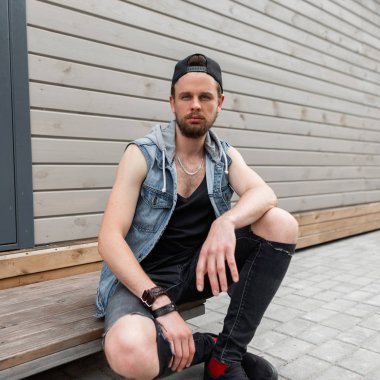 Bir şapka yırtık kot bir t-shirt bir kot yelek bir kap sakallı bir sakallı Brutal yakışıklı genç adam bir yaz günü bir vintage binanın yakınında ahşap bir adım üzerinde oturur. Amerikalı adam. Sokak stili. Erkek giyim