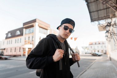 Şık bir sırt çantası ile güneş gözlüğü bir kap şık siyah giysiler içinde Yakışıklı genç hipster adam güneşli bir yaz parlak bir günde şehir etrafında yürüyor. Çekici kentsel adam hafta sonu sahiptir.