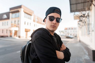 Bir sırt çantası ile şık güneş gözlüğü siyah bir sweatshirt şık bir kap yakışıklı genç Amerikalı hipster adam bir vintage binanın yakınında sokakta pozlar. Çekici adam şehir etrafında seyahat
