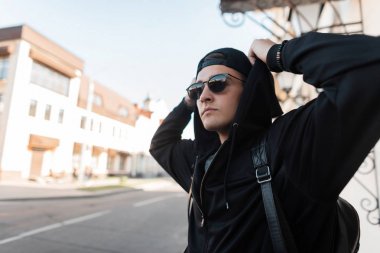 Bir sırt çantası ile güneş gözlüğü bir beyzbol şapkası bir kapüşonlu sweatshirt Yakışıklı genç hipster adam güneşli bir günde bir vintage binanın yakınında duruyor. Şehir sokaklarında seyahat eden Amerikalı adam.