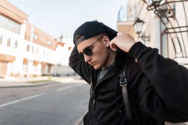Şık bir sırt çantası ile bir kap şık güneş gözlüğü ile şık bir sweatshirt Modern genç yakışıklı hipster adam binanın yakınında duruyor ve kaputu düzelter. Yürüyüş için çekici adam.