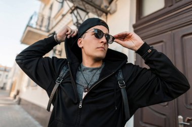 Şık bir sırt çantası ile güneş gözlüğü bir trendy kap şık siyah giysiler içinde Amerikalı genç hipster adam güneşli bir yaz gününde şehir etrafında yürüyor. Çekici kentsel adam modeli hafta sonu sahiptir.
