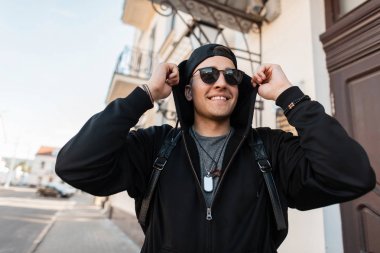 Bir kap bir sırt çantası ile şık bir sweatshirt şık güneş gözlüğü Pozitif yakışıklı genç hipster adam ayakta ve sokakta bir vintage binanın yakınında gülümsüyor. Neşeli adam açık havada.