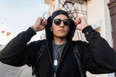 Bir moda sırt çantası ile bir kap vintage şık güneş gözlüğü ile şık bir sweatshirt Kentsel genç yakışıklı hipster adam binanın yakınında duruyor ve kaputu düzelter. Yürüyüş için serin adam.