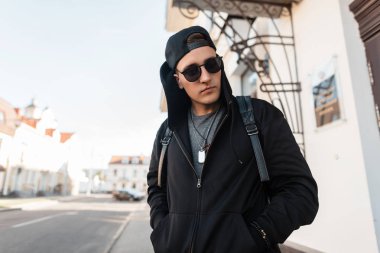 Bir sırt çantası ile şık güneş gözlüğü siyah bir sweatshirt şık bir kap güzel bir genç Amerikalı hipster adam bir vintage binanın yakınında sokakta pozlar. Çekici adam seyahat eder. Sokak stili.