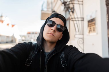 Şık güneş gözlüğü bir başlık ve binanın yakınındaki şehirde elinde bir kamera ile bir kap ile şık bir sweatshirt yakışıklı bir hipster adam Portresi. Yakışıklı adam açık havada selfie yapar.