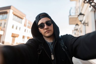 Şık kentsel genç adam hipster bir beyzbol şapkası bir trendy kapüşonlu sweatshirt vintage güneş gözlüğü güneşli bir günde sokakta selfie yapar. Çekici adam açık havada güneşin tadını çıkarır. Sokak modası.