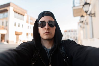 Siyah kapüşonlu şık bir kap ile trendy koyu güneş gözlüğü Ciddi yakışıklı genç adam hipster güneşli bir yaz gününde şehirde bir selfie yapar. Nice Amerikalı adam vintage binaların yakınında yürüyor. 