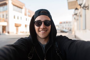 Sevimli bir gülümseme ile bir beyzbol şapkası siyah kapüşonlu şık güneş gözlüğü mutlu genç hipster adam elinde kamera tutan bir selfie yapar. Güneşli bir günde şehirde çekici neşeli adam.