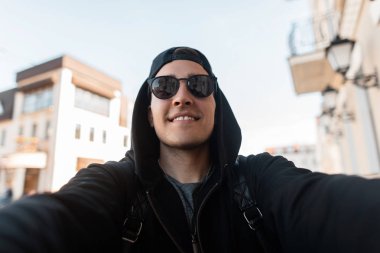 Şık güneş gözlüğü bir başlık ve binanın yakınındaki şehirde elinde bir kamera ile bir kap ile şık bir sweatshirt mutlu bir hipster adam Portresi. Neşeli adam açık havada selfie yapar.