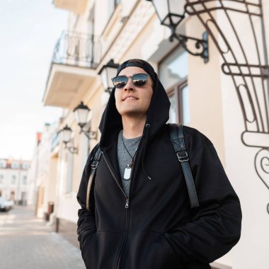 Güneşli bir yaz gününde, güneş gözlüğü takmış şık siyah elbiseli, sevimli gülümseyen genç hippi adam şehirde şık bir sırt çantası takıyor. Neşeli, çekici, kentli adam yürüyüşü seviyor.