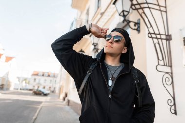 Şık bir kap güneş gözlüğü ile şık bir siyah kapüşonlu sweatshirt Neşeli genç hipster adam şehirde bir binanın yakınında yolun yanında duruyor. Şehir adam sokakta güneşli bir gün sahiptir.