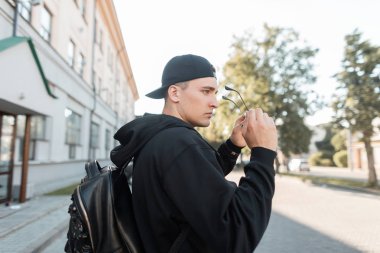 Bir sırt çantası ile güneş gözlüğü bir beyzbol şapkası bir kapüşonlu sweatshirt Kentsel genç hipster adam güneşli bir günde bir vintage binanın yakınında yürüyor. Amerikalı iyi adam şehir sokaklarında seyahat.
