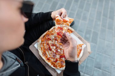 Lezzetli sıcak pizza bir dilim tutan güneş gözlüğü şık genç adam. Adam sokakta oturmuş pizza yiyor. Yemek vakti. Close.