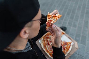 Lezzetli sıcak pizza açık bir dilim ile koyu güneş gözlüğü şık genç adam hipster. İyi adam sokakta oturmuş pizza yiyor. Yemek vakti. Close.