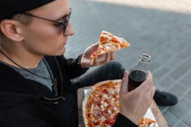 Güneş gözlüğü bir kap şık giysiler şık genç hipster adam pizza yiyor ve bir cam şişe bir içki içiyor sokakta oturuyor. Yakışıklı adam dışarıda öğle yemeği yiyor..