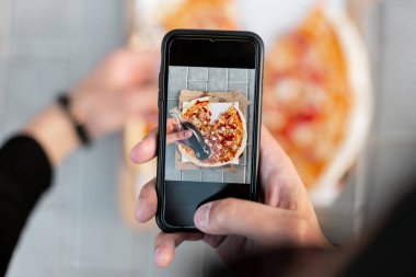 Genç adam akıllı telefondan lezzetli pizzaların fotoğrafını çekiyor. Ekranda erkek eli ve pizza görüntüsü var. Üst görünüm.