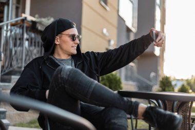Spor ayakkabı bir kapüşonlu siyah bir beyzbol şapkası koyu güneş gözlüğü neşeli hipster genç adam gülümsüyor ve bir yaz kafede bir şehirde otururken selfie alır. Hoş neşeli bir adam. Erkek giyim.