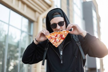 Bir kap bir başlık ile siyah moda sweatshirt trendy güneş gözlüğü Kentsel genç hipster adam şehirde bir sokakta yüzünün yanında pizza bir dilim tutar. Yakışıklı adam pizza ısırıyor