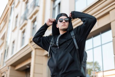 Modern genç yakışıklı hipster adam şık bir sweatshirt şık güneş gözlüğü ile bir kap ile şık bir sırt çantası binanın yakınında poz ve kaput doğrultma. Yürüyüş için çekici adam.