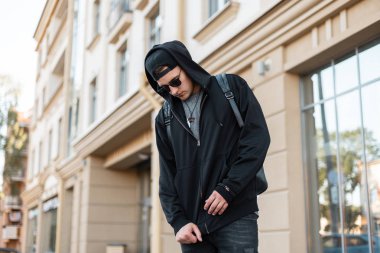 Bir sırt çantası ile bir kap moda güneş gözlüğü şık kapüşonlu sweatshirt Güzel genç hipster adam modern bir binanın yakınında bir şehirde açık havada duruyor. Çekici adam modeli sokakta yürür.