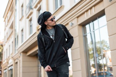 Şık bir sırt çantası ile güneş gözlüğü bir kap şık siyah giysiler içinde Yakışıklı genç hipster adam güneşli bir yaz şehirde yürüyor. Amerikalı çekici kentsel turist adam hafta sonu sahiptir
