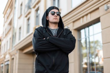 Güneş gözlüğü şık bir kap şık bir siyah kapüşonlu sweatshirt Çekici genç hipster adam parlak bir günde şehirde bir vintage binanın yakınında poz lar. Yakışıklı şehirli adam dışarıda. Moda.