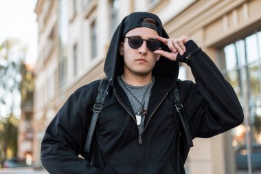 Bir sırt çantası ile siyah bir kap güneş gözlüğü bir başlık ile şık bir siyah sweatshirt Sevimli genç hipster güneşli bir yaz gününde şehir etrafında seyahat. Sokaktaki çekici adam. Erkek giyim.
