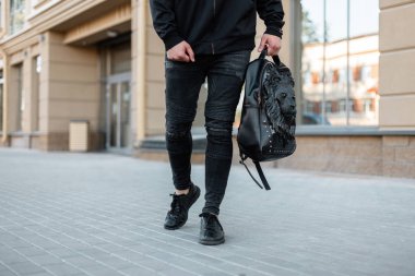 Bir aslan desenli bir deri sırt çantası ile vintage kot şık bir siyah sweatshirt şık genç adam binaların yakınındaki cadde boyunca yürüyor. Modern adam dışarıda. Close. Erkek giyim