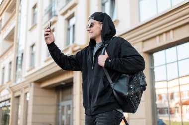 Sevimli bir gülümseme ile bir kap şık güneş gözlüğü kapüşonlu trendy kapüşonlu Mutlu yakışıklı genç hipster adam bir vintage binanın yakınında şehirde selfie yapar. Neşeli adam güneşli bir günün tadını çıkarır. Erkek giyim.