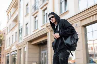 Kot şık bir sırt çantası ile siyah bir kap güneş gözlüğü ile trendy kapüşonlu sweatshirt trendy genç hipster adam parlak bir yaz gününde şehir etrafında seyahat. Sokakta çekici adam. 