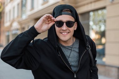 Şık güneş gözlüğü içinde mutlu yakışıklı genç hipster adam bir kap bir sırt çantası ile şık bir sweatshirt ayakta ve sokakta bir vintage binanın yakınında gülümsüyor. Neşeli adam açık havada.