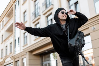 Siyah bir sweatshirt şık bir sırt çantası ile trendy güneş gözlüğü bir kap kentsel neşeli genç adam otostopçu şehirde duruyor ve bir taksi yakalamak için çalışıyor. Mutlu adam arabayı yakalamaya çalışıyor.