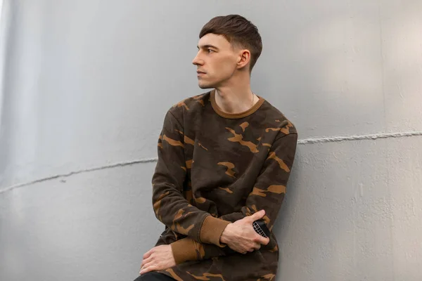 Jovem sério agradável em uma camisa militar verde elegante c penteado elegante repousa perto da parede cinza metal em um dia ensolarado de verão. Bonito modelo ao ar livre. Moda masculina . — Fotografia de Stock