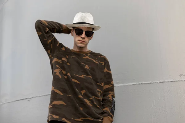 Hombre hipster americano de moda en un sombrero vintage de paja con gafas de sol oscuras en una camisa militar de moda posando cerca de un edificio de metal gris en la ciudad. Guapo chico moderno es relajante al aire libre . — Foto de Stock