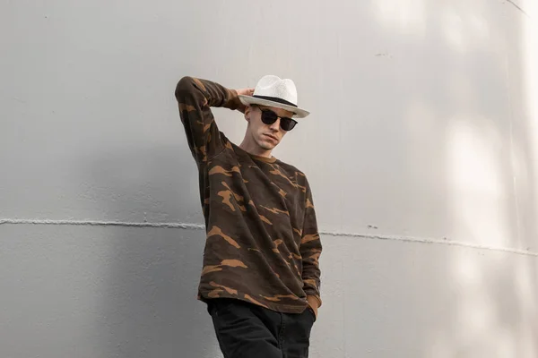 Homem hipster jovem bonito americano em uma camisa militar na moda em óculos de sol elegantes em um chapéu em jeans posa perto de uma parede cinza metálica ao ar livre. Cara atraente em uma caminhada em um dia ensolarado de verão . — Fotografia de Stock