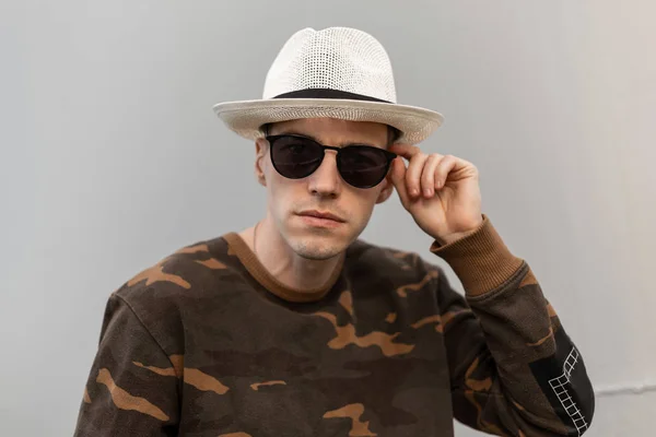 Modern handsome young hipster man in a vintage straw hat in a fashionable camouflage shirt straightens stylish sunglasses near a metal wall. Nice guy model posing outdoors. Trendy menswear. — Stock Photo, Image