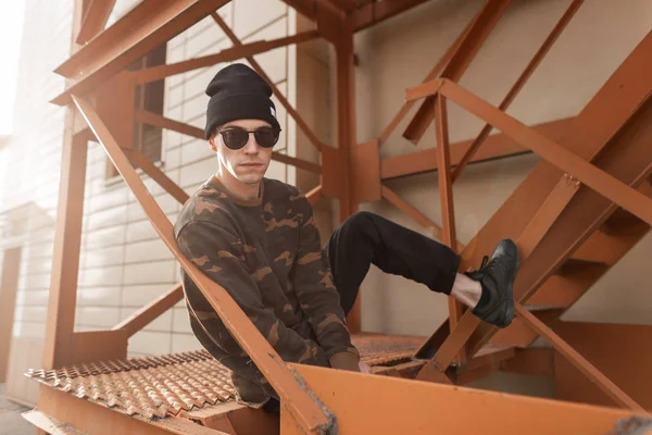 Askeri gömlekli, güneş gözlüklü, kot pantolonlu, güneş gözlüklü genç, yakışıklı hippi güneşli bir yaz gününde şehirdeki bir binanın yanındaki demir bir merdivende oturuyor. Havalı bir adam..