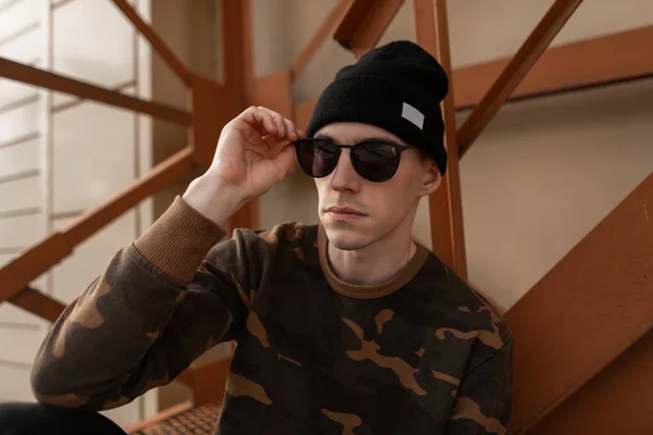 Guapo joven hipster hombre en gafas de sol de moda en un sombrero de punto negro en una camisa militar en jeans está sentado cerca de las escaleras de hierro naranja en la ciudad. Atractivo chico moderno está descansando en la calle . — Foto de Stock