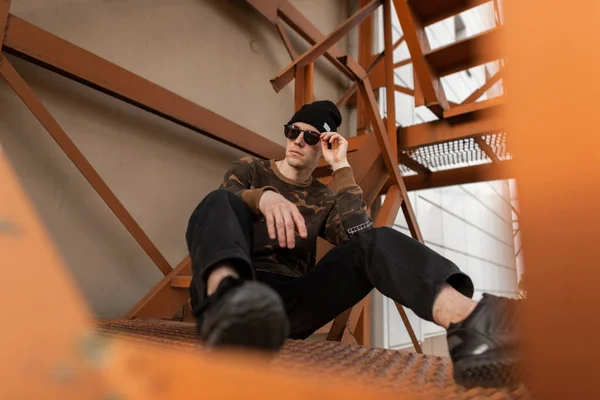 Κομψό νεαρό χίπστερ σε ένα στρατιωτικό πουκάμισο με τζιν σε μαύρο καπέλο σε σκούρα γυαλιά ηλίου σε αθλητικά παπούτσια ανάπαυσης που κάθονται στις μεταλλικές πορτοκαλί σκάλες στην πόλη. Ελκυστικός σύγχρονος τύπος. Το στυλ του δρόμου. Καλοκαίρι. — Φωτογραφία Αρχείου