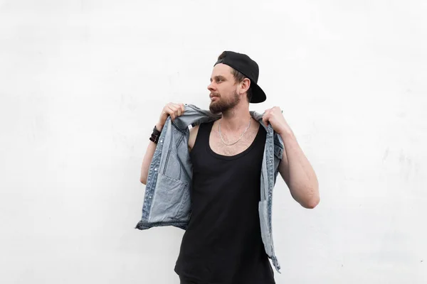 Homem jovem hipster atraente em colete de ganga elegante em camiseta preta em poses de boné na moda perto de parede vintage branco ao ar livre. Um tipo simpático na rua num dia de Verão. Roupas masculinas elegantes . — Fotografia de Stock