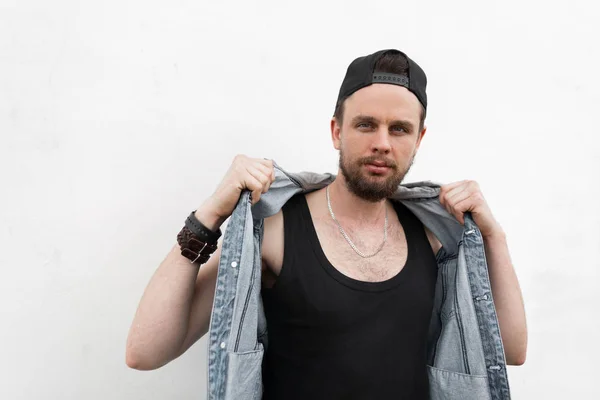 Young cool hipster man in a stylish black cap in a fashionable t-shirt stands near a white wall in the city and straightens blue denim vest. Nice handsome guy model posing. Modern men's street style. — Stock Photo, Image