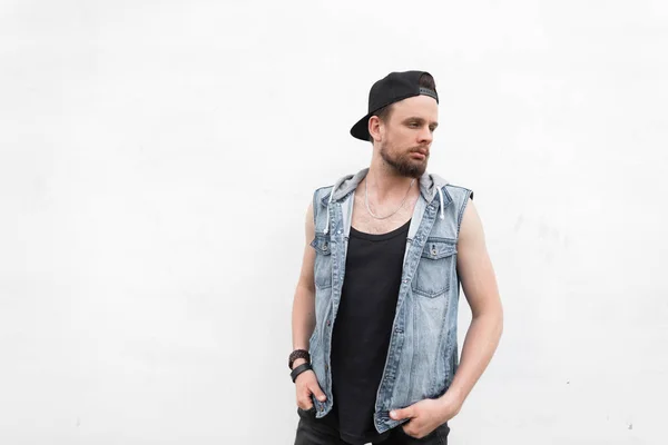 Jeune homme séduisant dans un T-shirt en gilet en jean dans un chapeau noir à la mode avec une barbe est debout près d'un mur vintage blanc un jour d'été lumineux. Beau mec en promenade dans la ville . — Photo